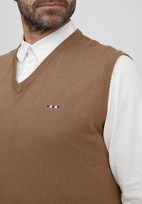 Brun stickad väst med V-halsdesign, bärs över en vit jockskjorta. Har en liten flerfärgad logotyp på bröstet.