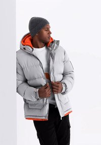 Veste en duvet grise avec doublure orange, capuche amovible et fermeture éclair. Portée par-dessus une chemise blanche et associée à un pantalon cargo noir.