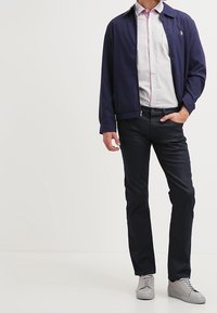 Man in een marineblauwe jas, lichtgestreept shirt, donkere broek en grijze sneakers, staand met één hand in de zak op een effen achtergrond.