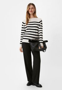 Maglione a righe bianco e nero con maniche lunghe, abbinato a pantaloni larghi neri. Una borsa di pelle nera è tenuta in una mano.