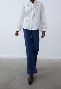Camicia bianca con chiusura a bottoni, maniche lunghe e cuciture a contrasto. Abbinata a pantaloni in denim a zampa d'elefante blu e stivaletti neri a punta.