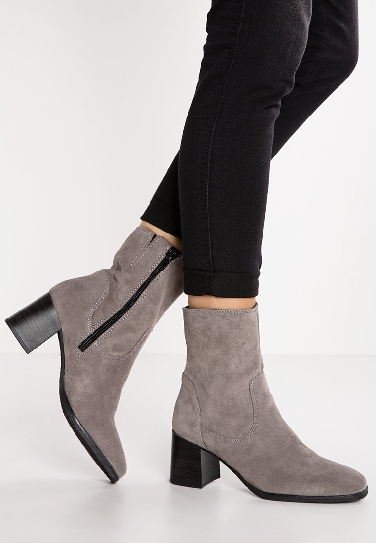 Personne portant des bottines en daim gris avec des talons blocs noirs et des fermetures éclair latérales, associées à un pantalon skinny noir retroussé.