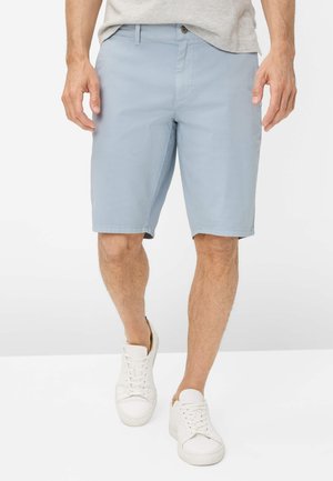 Mand iført lyseblå knælange shorts og hvide sneakers går på en ensfarvet hvid overflade.