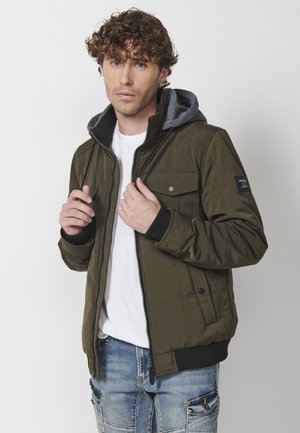 Olive green jacket with a gray hood, front zip closure, two chest pockets, ribbed cuffs, and stitched detailing on the fabric.