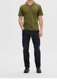 Polo à manches courtes vert olive avec un col plat, associé à un jean bleu foncé et des chaussures noires. Tissu simple et texturé avec une coupe décontractée.