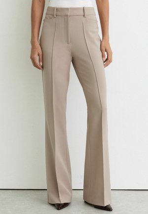 Kvinde iført beige skræddersyede bukser med frontsyninger, bæltestropper, sidelommer og brune spidse sko foran en ensfarvet hvid væg.