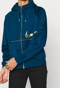 Person som bär en blå zip-up hoodie med en glänsande guldig Nike swoosh-logga på bröstet, i kombination med svarta byxor.