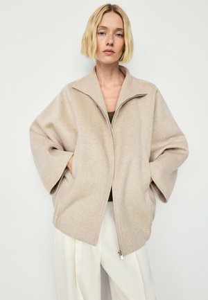 Frau mit kurzem blondem Haar trägt eine lockere beige Zip-Jacke über einem dunklen Oberteil und cremefarbene Hose, steht vor einem schlichten, hellen Hintergrund.