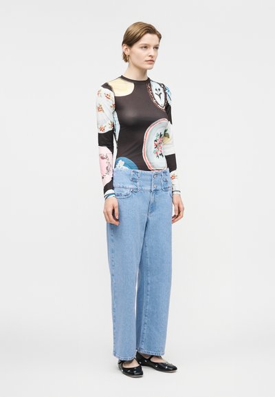 Γυναίκα στέκεται φορώντας πολύχρωμο μακρυμάνικο τοπ με λουλουδάτο μοτίβο, ανοιχτόχρωμο τζιν παντελόνι με φαρδιά μπατζάκια και μαύρα μπαλαρίνια με τρουκς.