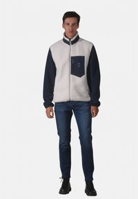 Jeune homme portant une veste polaire blanche et marine, un jean bleu foncé et des bottes noires à lacets, debout devant un fond uni.