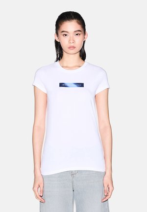 Mujer con el pelo liso y negro lleva una camiseta blanca de Armani Exchange y vaqueros azul claro, de pie frente a un fondo liso.