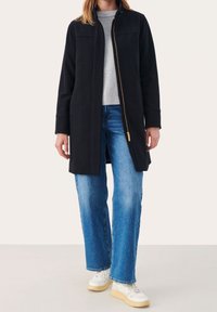 Sort lynlåsfrakke med en lige silhuet, der har en høj krave og frontlommer; bæres over en grå sweater og blå jeans.