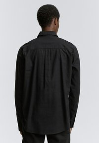 Zwarte langslijfs shirt van een gestructureerde stof. Heeft een klassieke kraag, met knopen sluitingen aan de mouwen, en een rechte zoom met subtiele plooien.