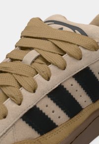 Μπεζ σουέτ αθλητικό παπούτσι με καφέ κορδόνια, τρεις μαύρες ρίγες και υφή στη σόλα. Διαθέτει μικρές οπές κατά μήκος των πλευρών.