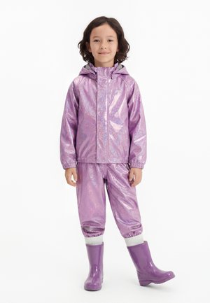 Bambino con capelli ricci scuri che indossa una giacca impermeabile viola lucido, pantaloni abbinati, calzini bianchi e stivali da pioggia viola lucidi, in piedi su sfondo bianco.