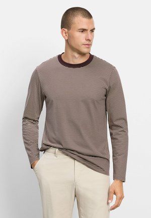 Uomo che indossa una camicia a righe marroni e bianche a maniche lunghe con pantaloni beige, guarda di lato con una mano in tasca.