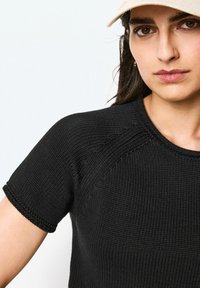 Svart kortärmad stickad topp med ribbat mönster, som har en rund halsringning och kontrasterande sömmar längs ärmarna.