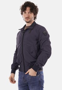 Mand med krøllet hår iført en mørk lynlåsbomberjakke og blå jeans, stående med den ene hånd i lommen mod en hvid baggrund.