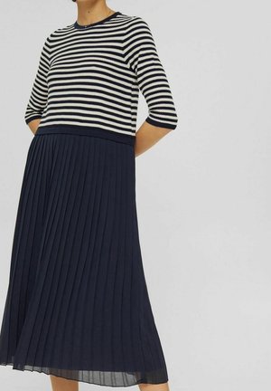 Mujer con falda midi plisada azul marino y camiseta de manga tres cuartos a rayas blancas y negras, de pie frente a un fondo claro y liso.