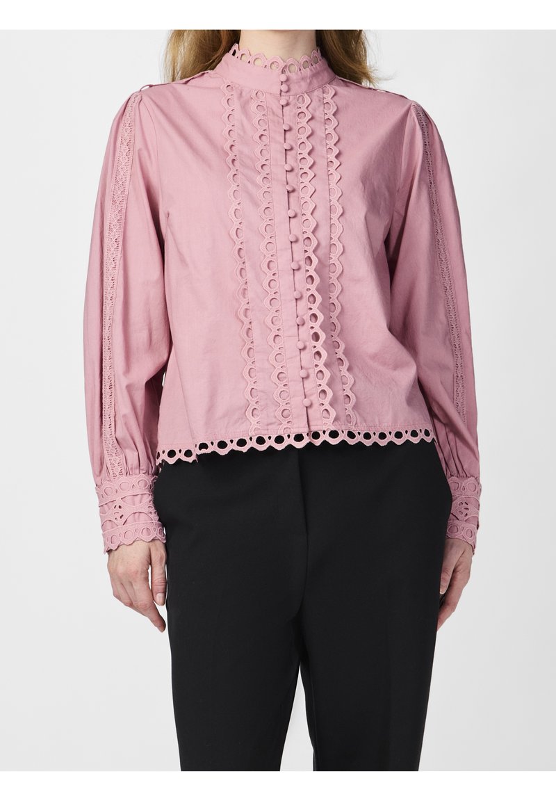 Femme portant un chemisier rose à manches longues avec bordure en dentelle, bords festonnés et boutons recouverts de tissu, associé à un pantalon noir.