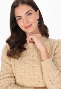 Maglione lavorato a maglia beige con motivo testurizzato, polsini a costine e scollatura rotonda. Il modello indossa orecchini circolari neri.
