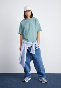 Tricou oversized mint cu un logo mic, pantaloni din denim albastri si o camasa cu dungi albastre deschise legata la talie. Sapca alba si adidasi.