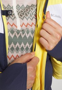 Giacca impermeabile Columbia, gialla con accenti blu navy, sopra un maglione a fantasia con colori verde, arancione e crema in motivi a diamante.