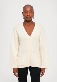 Kobieta z ogoloną głową, nosząca złote kolczyki w kształcie kół, beżowy sweter zapinany na guziki z kieszeniami oraz czarne spodnie, stojąca na jednolitym tle.