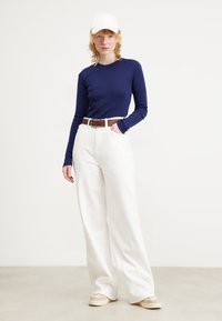 Top de manga larga azul marino, pantalones anchos blancos de cintura alta, cinturón marrón y zapatillas beige. Los accesorios incluyen una gorra blanca. Texturas suaves.