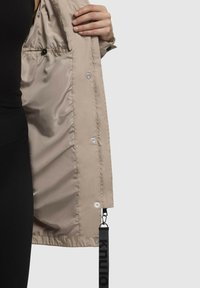 Giacca leggera beige con interno lucido, dotata di chiusure a pressione e una fascetta in tessuto con logo all'orlo. Design semplice e texture liscia.
