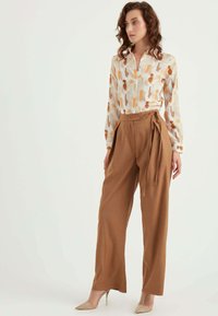 Bluse mit floralem Muster in hellem Creme mit orange und braunen Streifen, kombiniert mit hoch taillierten, schmalen braunen Hosen und nude-farbenen High Heels.