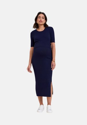 Schwangere Frau trägt ein marineblaues, geripptes Strick-Midikleid mit kurzen Ärmeln und seitlichem Schlitz, kombiniert mit weißen Sneakers vor einfarbigem Hintergrund.