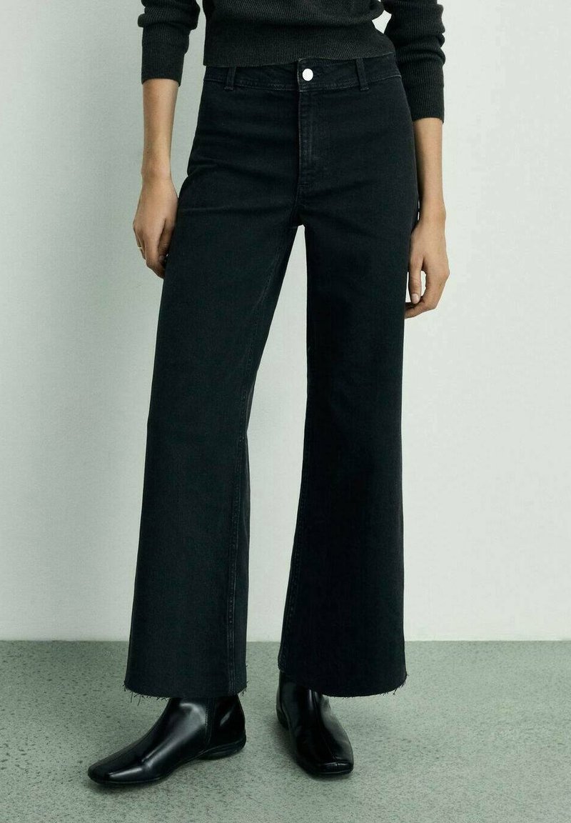 Personne portant un jean noir taille haute à jambes larges avec des ourlets bruts et des bottines noires contre un mur clair uni.