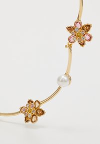 Collier floral doré avec des pierres roses et jaunes, présentant une perle blanche centrale. Métal lisse et détails complexes des pétales.