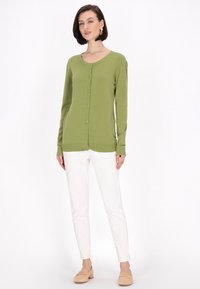 Cardigan verde con scollatura rotonda, maniche lunghe e bottoni frontali. Abbinato a pantaloni bianchi slim-fit e scarpe beige senza lacci.