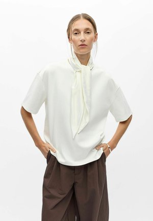 Femme aux cheveux clairs portant un haut blanc à manches courtes avec une écharpe assortie, un pantalon large marron et des bijoux minimalistes, les mains dans les poches.