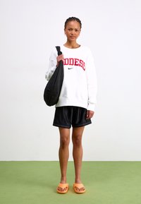 White sweatshirt with red "GODDESS" text, black shorts, orange slip-on sandals, and a black quilted shoulder bag. Standing on green floor.