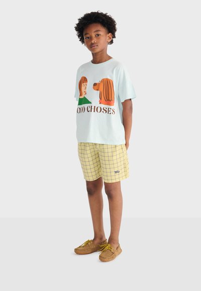 Chłopiec w jasnoniebieskiej koszulce z nadrukiem kreskówkowej postaci i psa oraz napisem "OSO CHOSES", żółtych szortach w kratę i beżowych mokasynach.