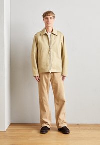 Veste légère beige avec col et fermeture éclair à l'avant, associée à un pantalon en velours côtelé marron clair et des chaussures foncées, debout sur un sol en bois.