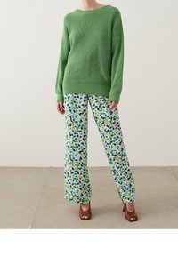 Grüner, gestrickter Pullover mit lockerer Passform, kombiniert mit weiten, gemusterten Hosen, die grüne, blaue und beige Sprenkel auf weißem Hintergrund aufweisen.