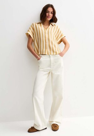 Kobieta stoi przy białej ścianie, ubrana w musztardowo-białą koszulkę w paski z krótkim rękawem, białe, szerokie spodnie oraz brązowe mokasyny.