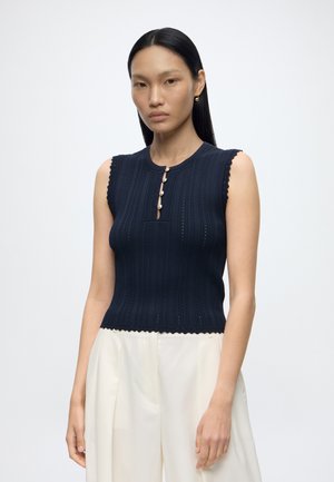Femme aux longs cheveux noirs portant un haut en maille bleu marine sans manches avec des boutons et un pantalon blanc taille haute sur fond uni.