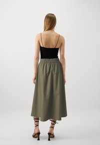 Schwarzes Oberteil mit Spitzenrücken, kombiniert mit einem fließenden olivgrünen Midirock, der einen elastischen Bund und schwarze Riemchen-Pumps hat.