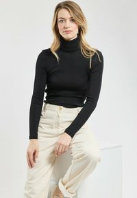 Frau mit blonden Haaren, die auf einem weißen Block sitzt und einen schwarzen Rollkragenpullover, beigetöne Hosen und weiße Sneaker trägt, vor einem schlichten Hintergrund.