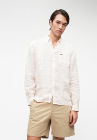 Chemise à manches longues rayée rose clair et blanche, boutonnée, avec une poche arborant un petit logo vert, accompagnée de shorts beiges.