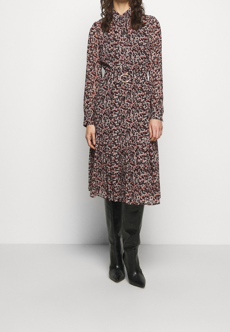 Femme portant une robe midi à manches longues à imprimé floral, avec une ceinture à la taille et des bottes noires montantes, debout devant un fond uni.