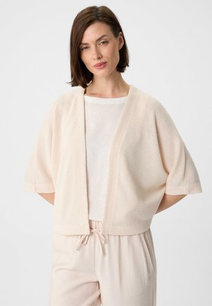 Helle rosa gestrickte Strickjacke mit kurzen Ärmeln, offener Vorderseite und lockerer Passform, kombiniert mit einem cremefarbenen Oberteil und sanften, beigen Hosen.