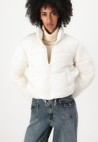 Giacca trapuntata bianca con colletto alto e dettagli in strass. Chiusura con zip frontale e orlo regolabile con cordino, abbinata a jeans a vita alta.