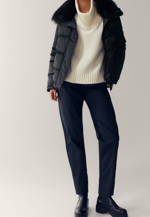 Personne portant une doudoune noire avec col en fourrure, un pull à col roulé blanc, un pantalon noir et des bottines noires, debout devant un fond uni.
