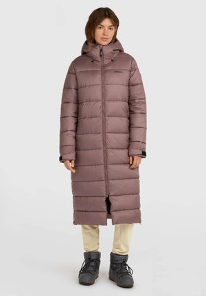 Frau trägt einen langen mauvefarbenen Daunenmantel mit Kapuze, beige Hose und schwarze Winterstiefel, steht vor einem schlichten hellen Hintergrund.